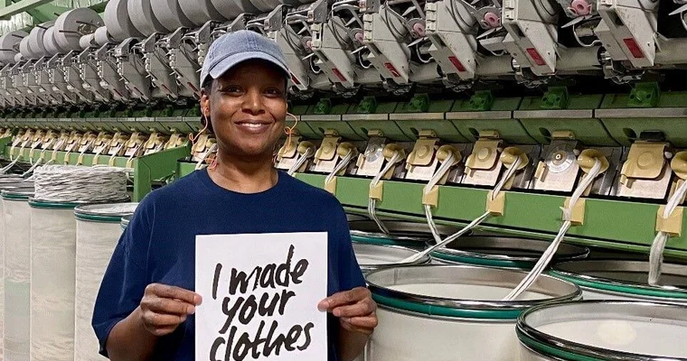 Jennifer tient une pancarte sur laquelle on peut lire « I Made Your Clothes » et sourit à la caméra.