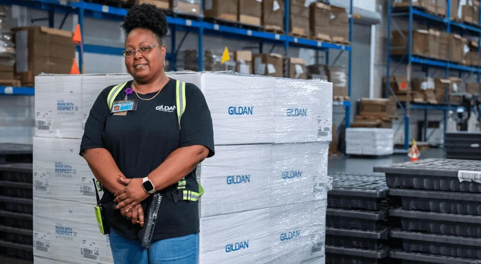 Une femme vêtue d'une chemise noire Gildan se tient à côté d'une pile de boîtes de distribution Gild